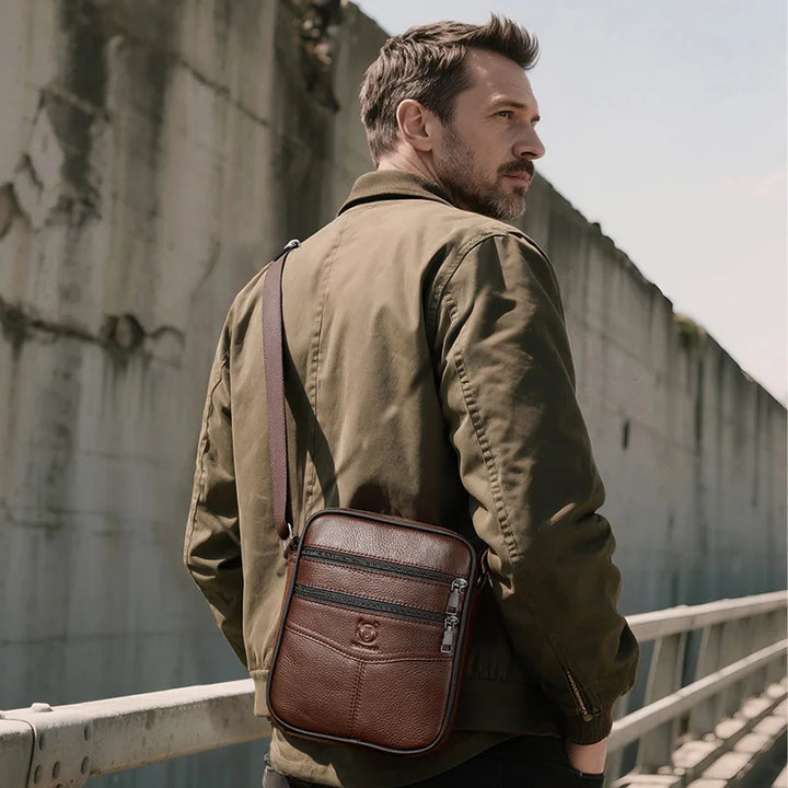 Man wearing a brown leather crossbody bag against a concrete wall.
