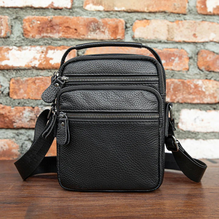 Black leather crossbody bag on a wooden surface with a brick wall background