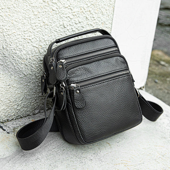 Black leather crossbody bag with multiple zippers on a textured surface.